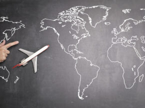 Top view of miniature airplane placed on over gray world map with crop hand of anonymous person indicating direction representing travel concept