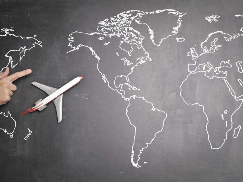 Top view of miniature airplane placed on over gray world map with crop hand of anonymous person indicating direction representing travel concept