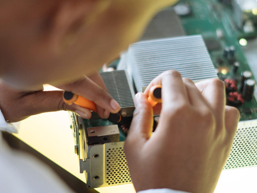 Crop of ethnic system administrator in uniform using screwdrivers and checking motherboard on electronic equipment during work in repair department